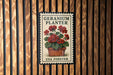 A vintage-style postage stamp featuring a potted geranium plant, mounted on a wooden wall.