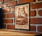 A wooden table with a brick wall in the background, and a framed stamp-like image of a hamster driving a car on the table.