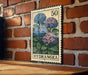 A vintage-style metal stamp with a floral design is displayed on a brick wall, featuring a close-up of three blue hydrangea flowers.