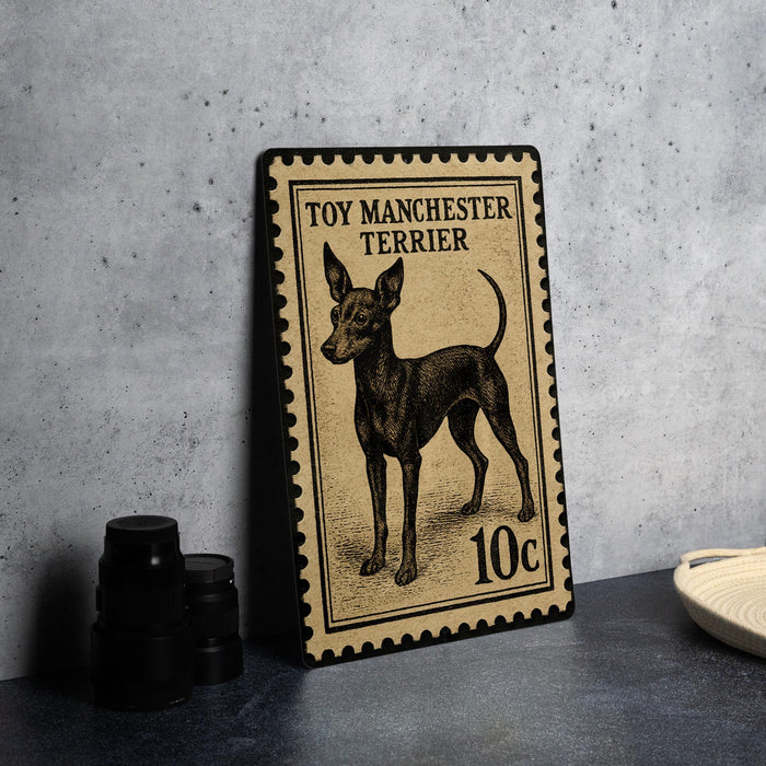 A toy Manchester Terrier dog stamp is displayed on a table, with a stack of black camera lenses and a white dish visible in the background.
