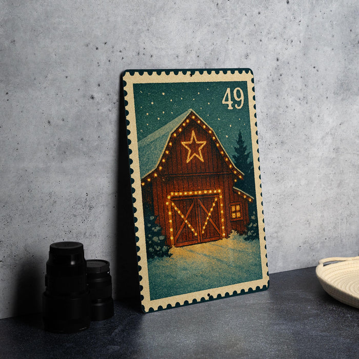 A metal stamp depicting a barn with a star on the roof, surrounded by lights and a snowy landscape, is placed on a gray countertop next to some black camera lenses.