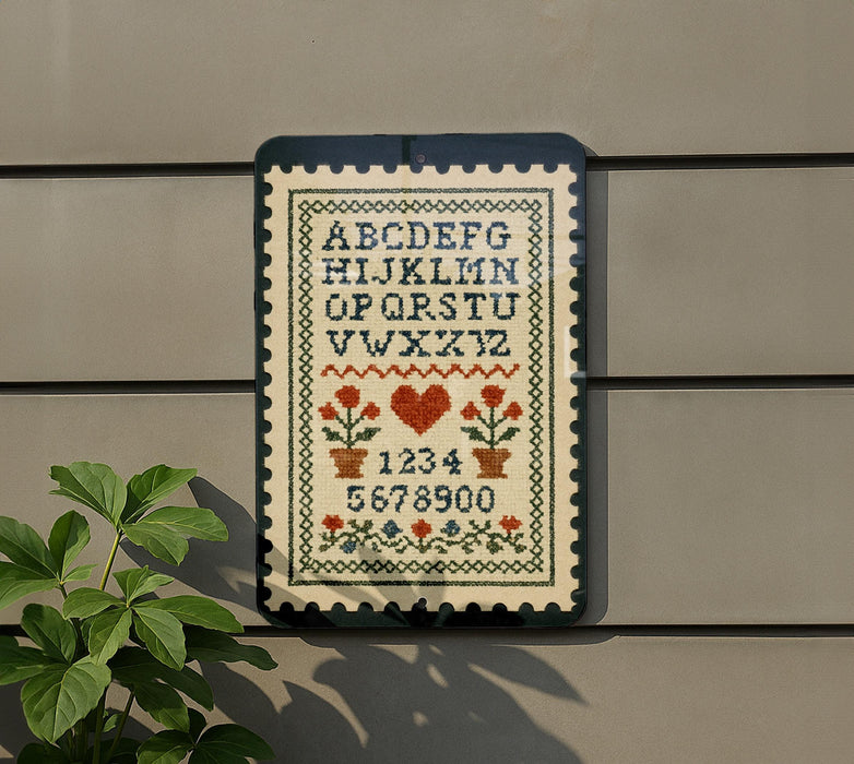 a cross-stitch pattern with the alphabet and numbers, hanging on a wall next to a plant.