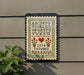 a cross-stitch pattern with the alphabet and numbers, hanging on a wall next to a plant.