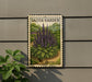 A framed print of a purple flower is hanging on a wall, with a plant to the left of it.