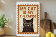 a sign with a black cat wearing glasses and the text "MY CAT IS MY THERAPIST" on it, placed on a wooden surface next to a potted plant and a cutting board.