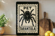A framed sign with a black and white illustration of a tarantula, accompanied by the text "PET TARANTULA" on a wooden shelf.