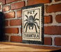 A metal sign with a black and white illustration of a tarantula, mounted on a brick wall.