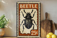 A vintage-style metal sign featuring a detailed illustration of a beetle, with the word "BEETLE" prominently displayed. The sign is placed on a wooden surface, with a potted plant and a wooden cutting board visible in the background.