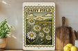 a vintage-style metal sign with a floral illustration of a field of daisies, placed on a wooden surface next to a wooden cutting board and some lemons.