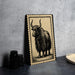 a framed illustration of a yak with long horns, placed on a table next to some black camera lenses.