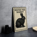 a silver fox rabbit illustration on a rectangular sign with a black border, placed on a gray countertop next to some black camera lenses.