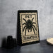 A framed poster of a tarantula is displayed on a countertop, with a stack of black camera lenses and a woven basket nearby.
