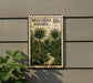 A metal sign with a tropical garden scene, featuring palm trees and a winding path, mounted on a wall.
