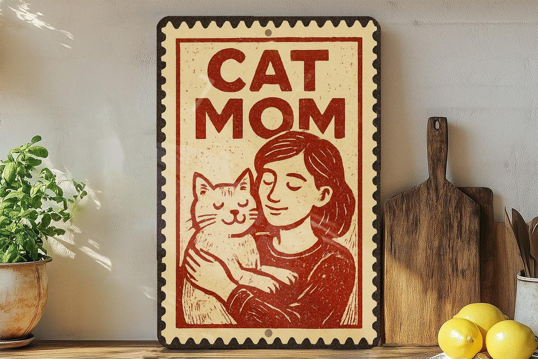 A vintage-style sign with the text "CAT MOM" and an illustration of a woman holding a cat, placed on a wooden surface next to a potted plant and a wooden cutting board.