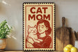 A vintage-style sign with the text "CAT MOM" and an illustration of a woman holding a cat, placed on a wooden surface next to a potted plant and a wooden cutting board.