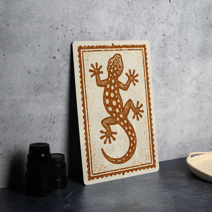 a decorative sign with a lizard design on a white background, placed on a dark surface next to some black objects.