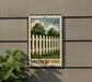 A white picket fence painted on a building, with a USA 24 postage stamp featuring a similar image of a white picket fence below it.