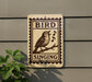 a sign that reads "BIRD SINGING" with a bird illustration on it. The sign is mounted on a wall, and there is a plant to the left of the sign.