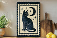 A framed illustration of a black cat sitting under a crescent moon and stars on a wooden shelf, accompanied by a wooden cutting board and a potted plant.