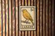A vintage-style postage stamp featuring a yellow canary perched on a branch, mounted on a wooden wall.