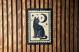 A black cat sitting on its hind legs with a crescent moon and stars above it, depicted on a wooden surface.