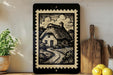 a framed illustration of a quaint cottage with a winding path, surrounded by a lush garden, on a shelf next to a wooden cutting board and a potted plant.