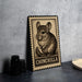 A framed illustration of a chinchilla is displayed on a table, with a stack of black camera lenses and a white plate visible in the background.