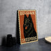 A metal sign featuring a black Belgian sheepdog with the text "BELGIAN SHEEPDOG" on it, placed on a countertop next to some black camera lenses.