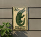 A green and white postage stamp featuring a crocodile, mounted on a wall next to a plant.