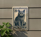 A 20-cent postage stamp featuring a cat, mounted on a wall next to a plant.