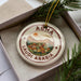 A decorative ornament in a box, featuring a circular design with a mountain landscape and buildings, along with the text "ABHA SAUDI ARABIA".