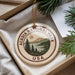 A circular ornament with the text "ACADIA NATIONAL PARK USA" and a scenic landscape design, hanging from a string.