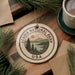 A circular ornament with the text "ACADIA NATIONAL PARK USA" and a scenic landscape design hangs on a wooden surface, surrounded by pine branches and a cup of coffee.