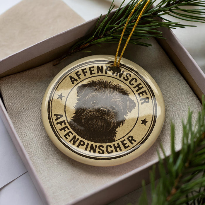 A circular ornament with a dog image and the text "Affenpinscheraffen" (Affenpinscher) on it, placed inside a box with pine branches.