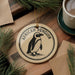 A decorative ornament featuring an illustration of an African penguin is displayed on a wooden surface, accompanied by a cup of coffee and pine branches.