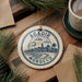 A decorative ornament with the text "AGADIR MOROCCO" and a graphic of a city skyline is displayed on a wooden surface, surrounded by pine branches and a cup of coffee.