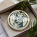 A ceramic ornament in a box, featuring a black and white illustration of a dog's face with the text "AKITA DOG" around it.