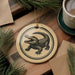a round ornament with a black and white illustration of a crocodile on a wooden surface, surrounded by pine branches and a cup of coffee.