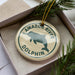 A round ornament with the text "AMAZON RIVER DOLPHIN" printed on it, surrounded by a green border. The ornament is placed inside a box with pine branches.