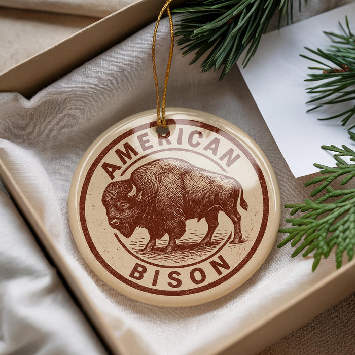 A circular ornament with the text "AMERICAN BISON" printed on it, featuring an illustration of a bison. The ornament is hanging from a string and is placed on a white surface, with some pine branches visible in the background.