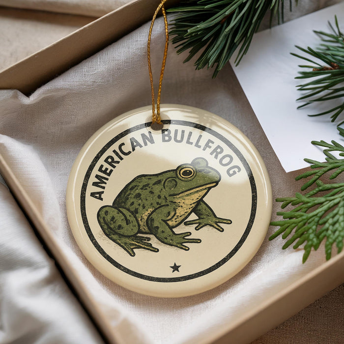 A circular ornament featuring a green frog, with the text "AMERICAN BULLFROG" printed on it, is displayed in a box with pine branches.