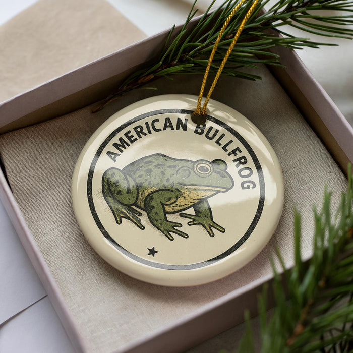 A circular ornament featuring a green frog, with the text "AMERICAN BULLFROG" printed on it, is displayed in a box surrounded by pine branches.