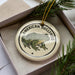 A circular ornament featuring a green frog, with the text "AMERICAN BULLFROG" printed on it, is displayed in a box surrounded by pine branches.