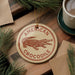 A circular ornament featuring an illustration of a crocodile is displayed on a wooden surface, surrounded by pine branches and a cup of coffee.