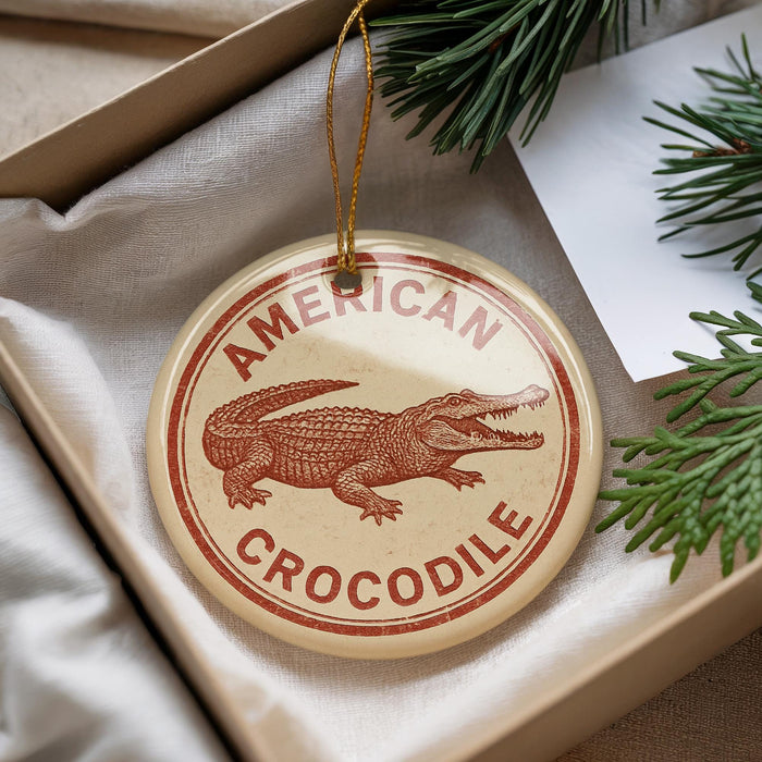 A circular ornament with the text "AMERICAN CROCODILE" printed on it, featuring an illustration of a crocodile. The ornament is hanging from a string and is placed inside a box with pine branches visible in the background.