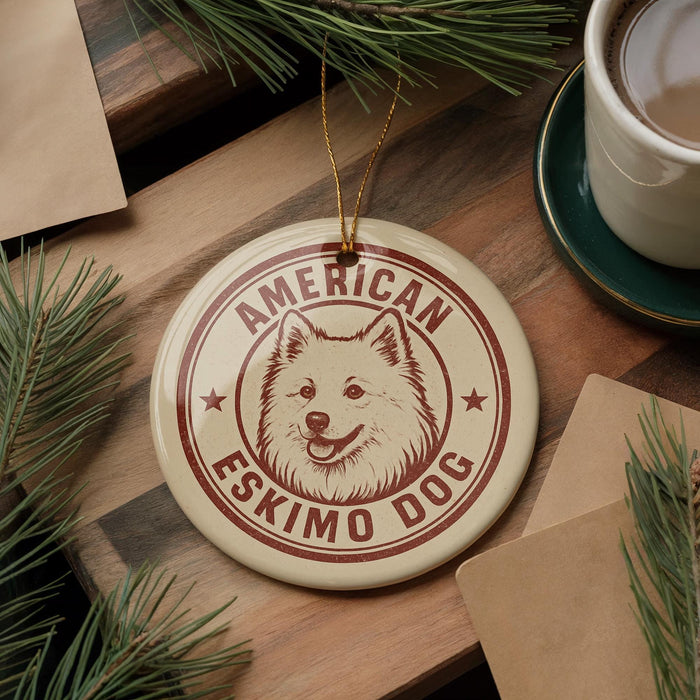 A circular ornament featuring an image of an Eskimo dog, with the text "AMERICAN ESKIMO DOG" printed on it, is displayed on a wooden surface alongside a cup of coffee and some pine branches.