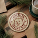 A circular ornament featuring an image of an Eskimo dog, with the text "AMERICAN ESKIMO DOG" printed on it, is displayed on a wooden surface alongside a cup of coffee and some pine branches.