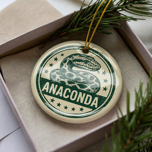 A circular ornament with a snake design and the word "ANACONDA" printed on it, placed inside a box with pine branches.