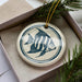 A ceramic ornament in the shape of a fish is displayed in a box, surrounded by pine branches.