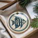 A decorative ornament featuring a blue and white striped fish is displayed in a box, surrounded by pine branches.